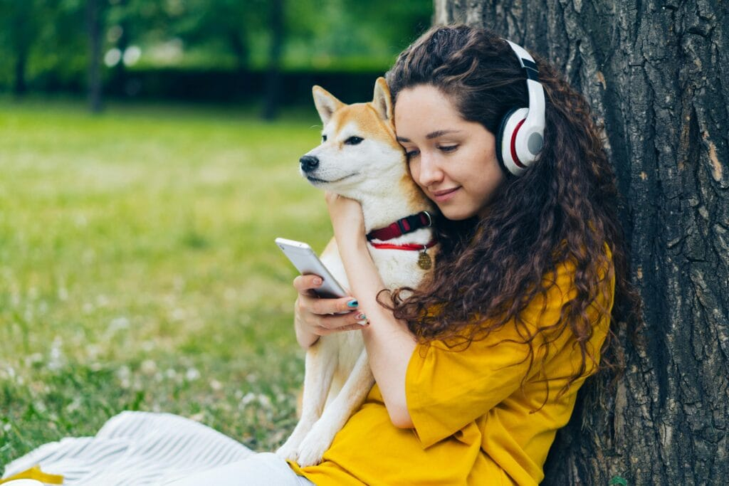 A woman in yellow cuddles a Shiba Inu while using a smartphone outdoors, enjoying music.