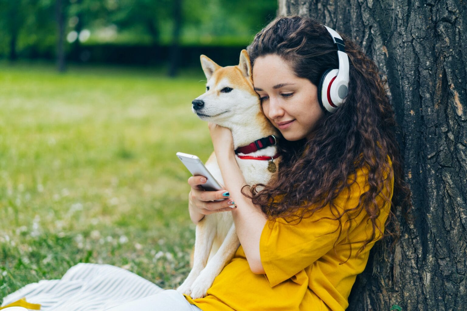 A woman in yellow cuddles a Shiba Inu while using a smartphone outdoors, enjoying music.