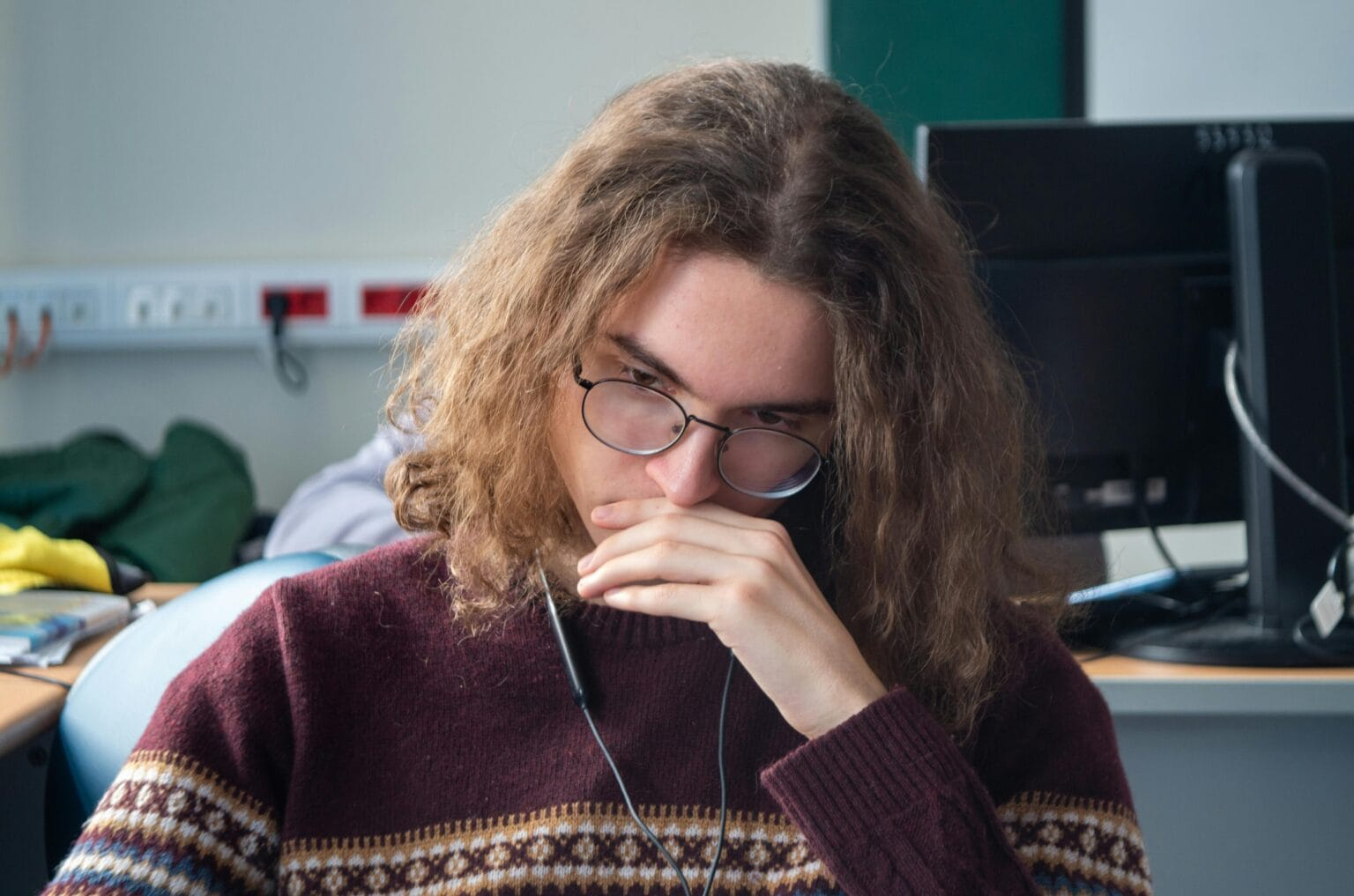 Young adult wearing glasses and sweater, deep in thought at a workstation.